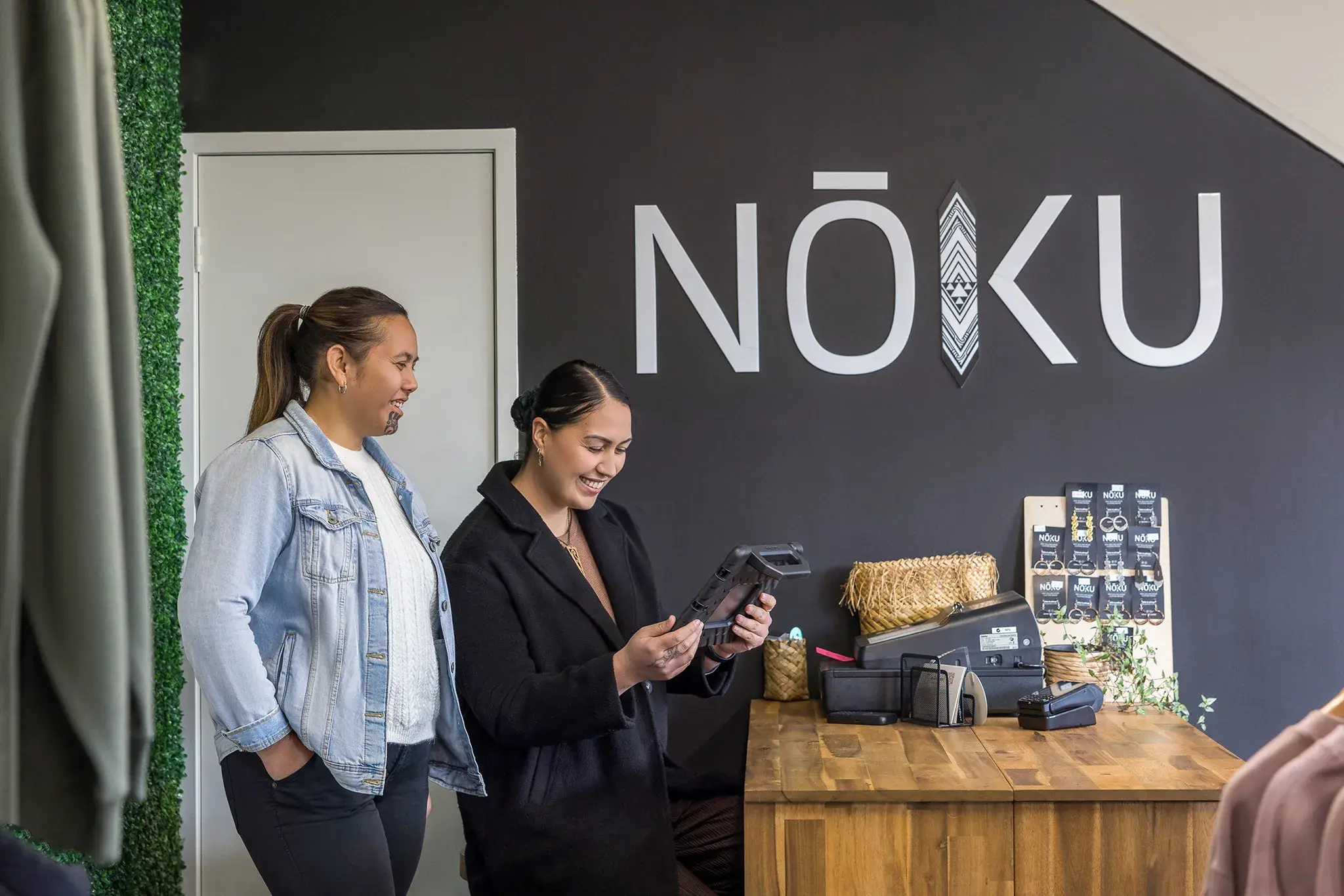 Two women inside the Nōku clothing store, one holding a tablet, illustrating the reliable broadband and local support provided by Now NZ to support Nōku's online and physical stores.