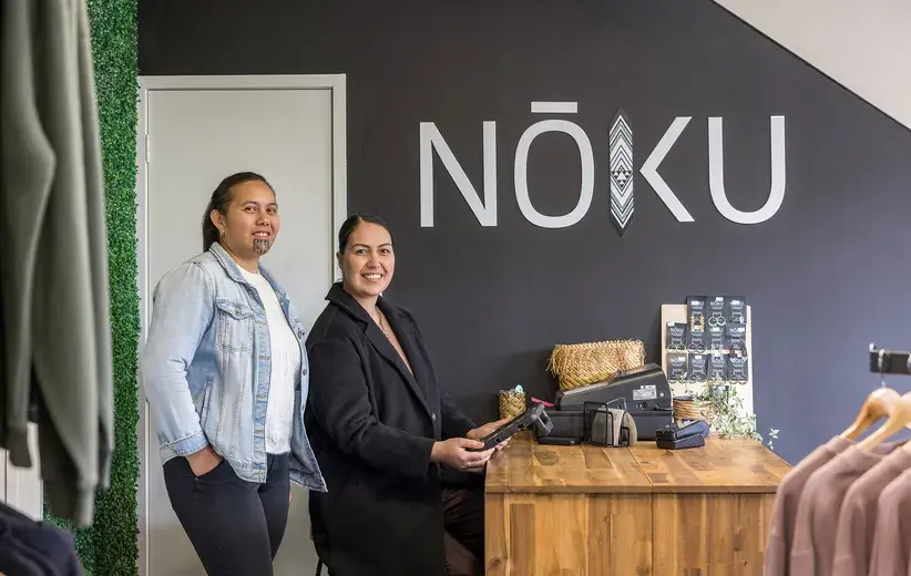 Two women in a store with the sign 'NŌKU' on the wall. Strengthen community connections through the facilitation of meaningful korero. Weaving technology with tradition, use Now's voice and broadband systems to support your kaupapa.