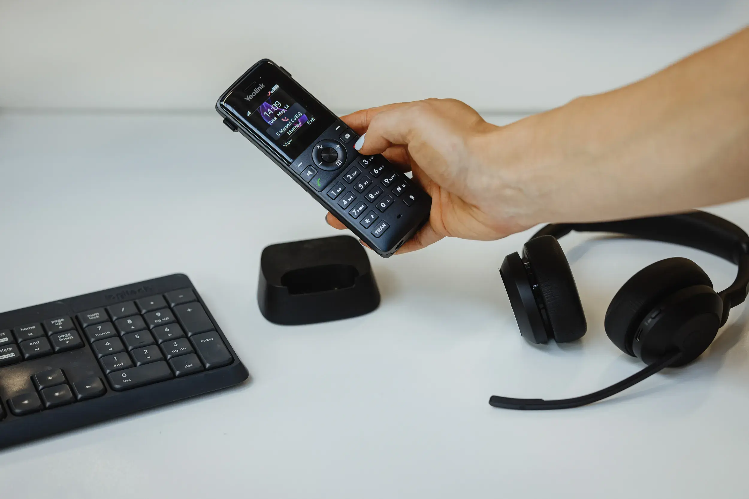 Person holding a cordless phone with a headset and keyboard nearby, representing the affordable business phone line feature bundle