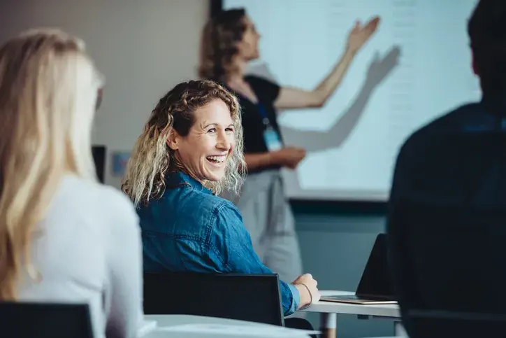 Smiling female educator attending a training session with a colleague presenting in the background, illustrating Now NZ's connectivity solutions and tech support for teaching and training professionals.