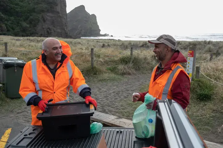 Two workers in orange safety gear having a conversation while standing near the beach, representing Now NZ's support in amplifying charity messages through effective business communication solutions.