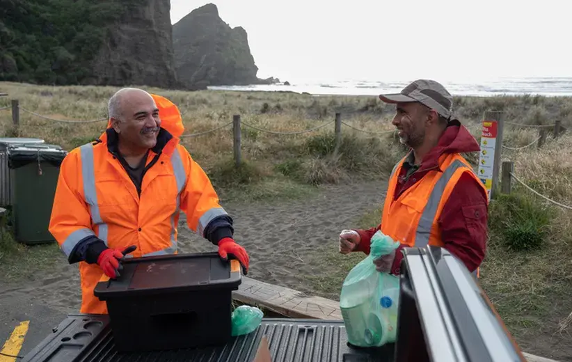 Two charity workers in bright orange jackets smiling and talking while organizing supplies at a coastal location, illustrating Now NZ's reliable telco solutions for charities to focus on making an impact.