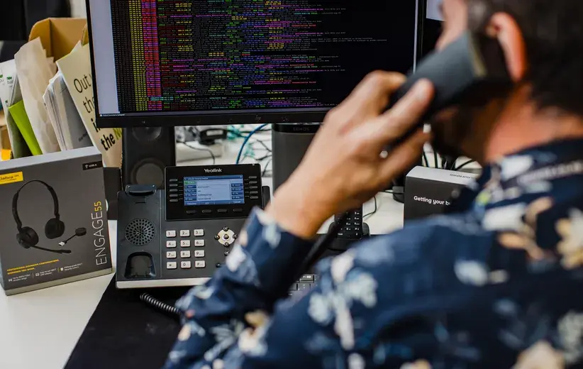 A man talking on a phone in front of a computer displaying code. Minimise your overheads without compromising on quality. Our telecommunications solutions are tailored to meet your practical and financial needs, offering the best of both worlds.