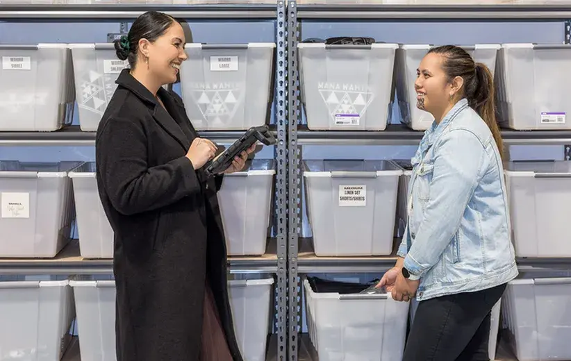 Two women working in a storage area with labeled bins, one holding a tablet, showcasing Now NZ's phone products supporting efficient business operations.