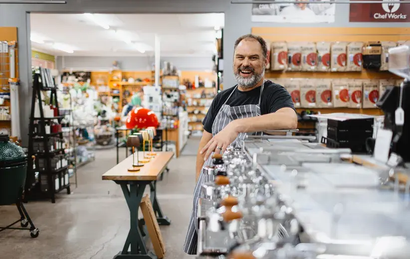 A cheerful store owner wearing an apron, standing beside a row of coffee machines in a well-stocked store, illustrating Now NZ's broadband and internet services that support local businesses.