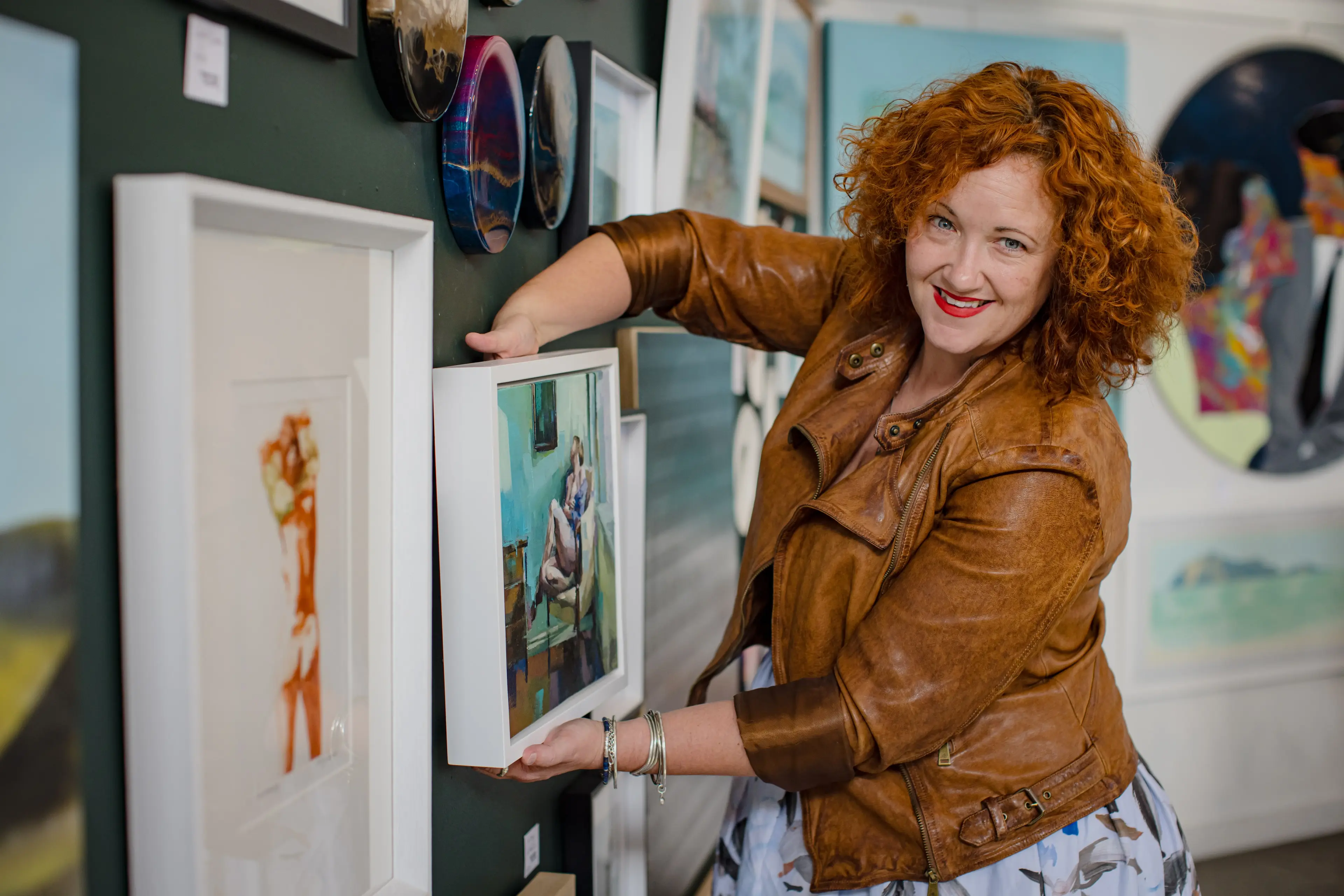 Smiling woman in a brown leather jacket arranging artwork on a wall, representing the ease of customer connections with Now's toll-free solutions