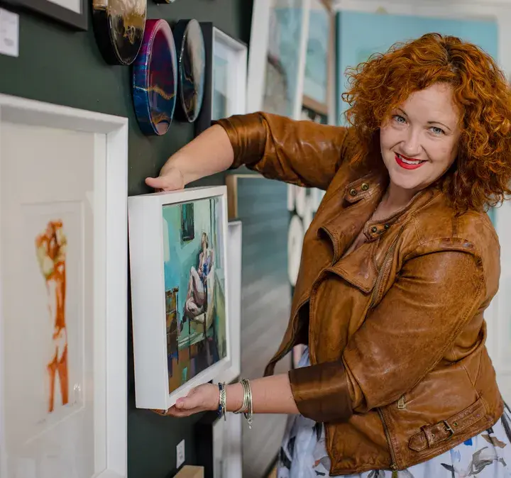 Smiling woman in a brown leather jacket arranging artwork on a wall, showcasing the reach and accessibility Now Reach provides to New Zealand customers