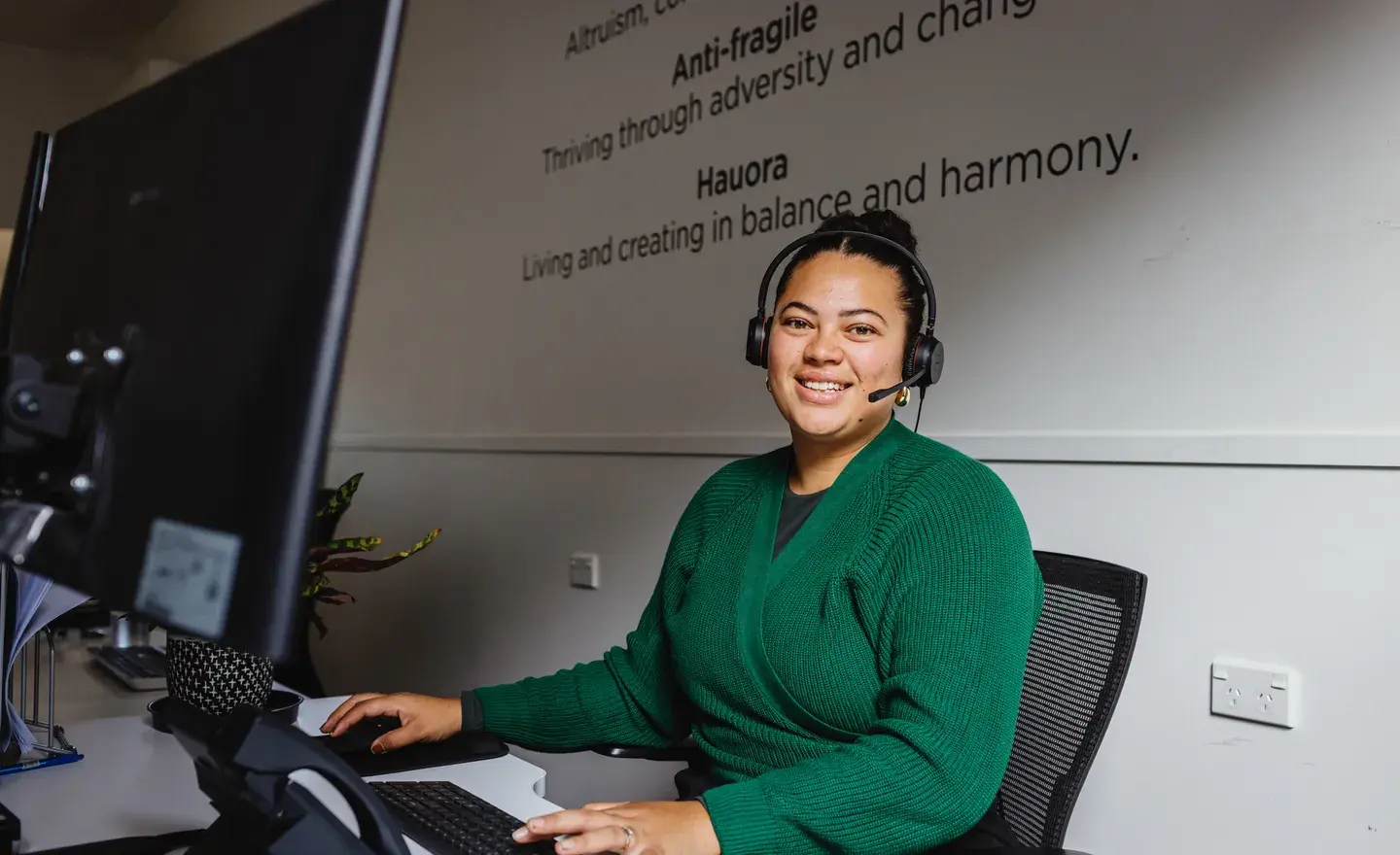 A smiling woman wearing a headset and a green sweater sits at her desk in a modern office, highlighting Now's contact centre solutions. Now offers a range of options from simple call distribution to complex contact centres, tailored to meet the needs and budget of your business.