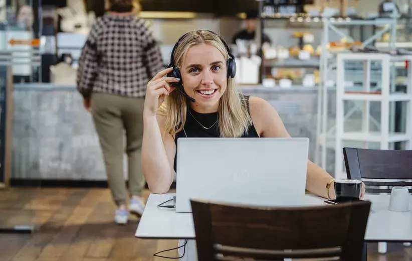 A woman working on a laptop in a café, wearing a headset and smiling, representing Now's telco technology solutions for sole traders to stay connected and maintain productivity.