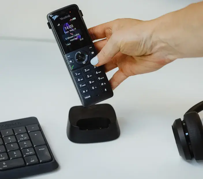 A hand placing a cordless office phone in its charging dock, accompanied by a keyboard and headphones, symbolizing Now NZ's commitment to keeping charities connected with reduced rates on essential tools.