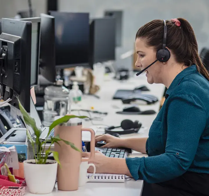 A woman wearing a headset working at her desk in a busy office environment, highlighting Now’s Mitel Contact Centre service. The service offers customisation to meet organisational needs, advanced call centre features such as call categorisation, place-in-queue announcements, CRM integration, and advanced reporting, backed by Mitel and in-house experts.