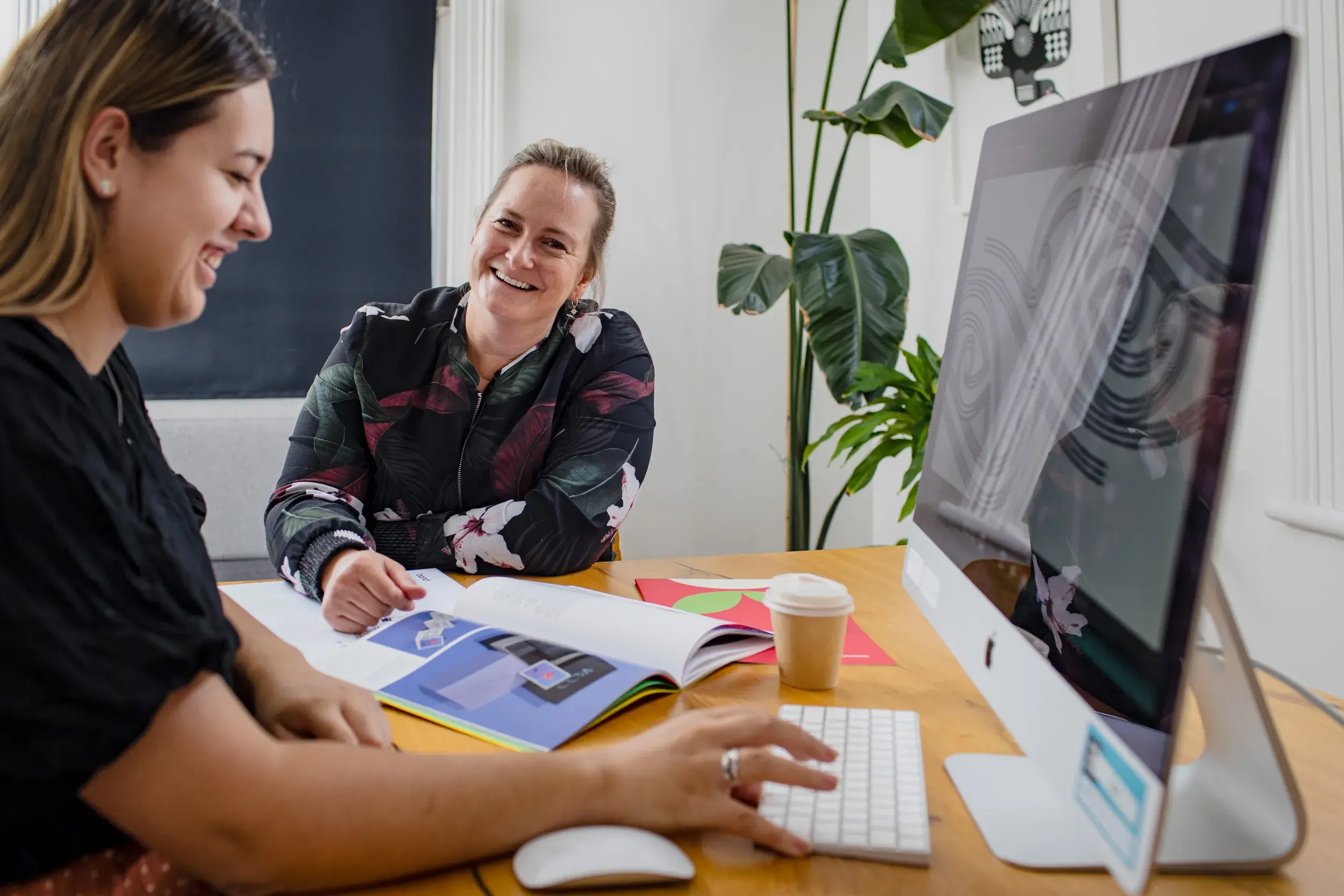 Two women smiling and working on a computer at a desk, with a book open and a coffee cup nearby