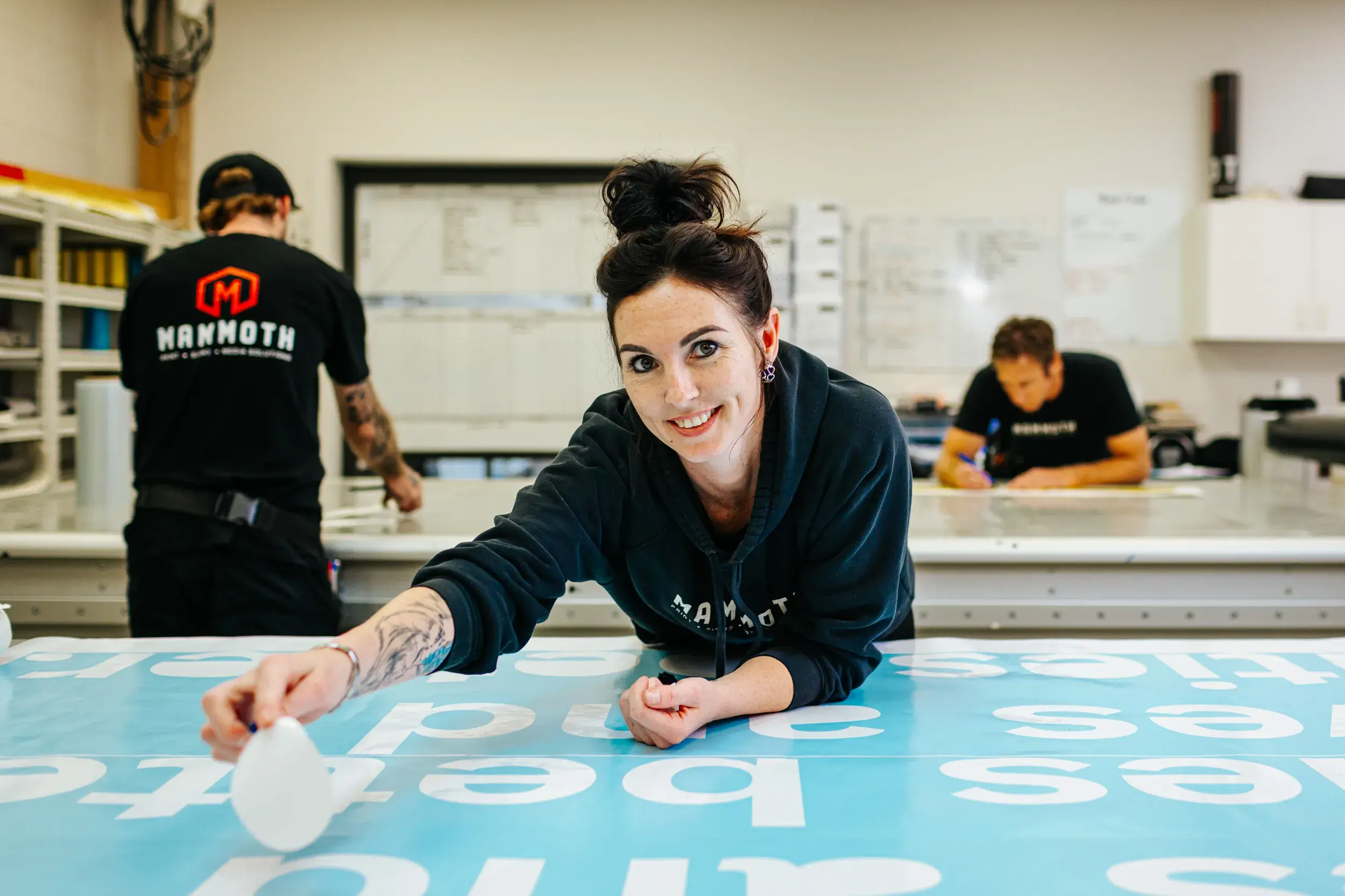 Female print shop staff member smiling while smoothing out a large printed sheet, with two colleagues working in the background