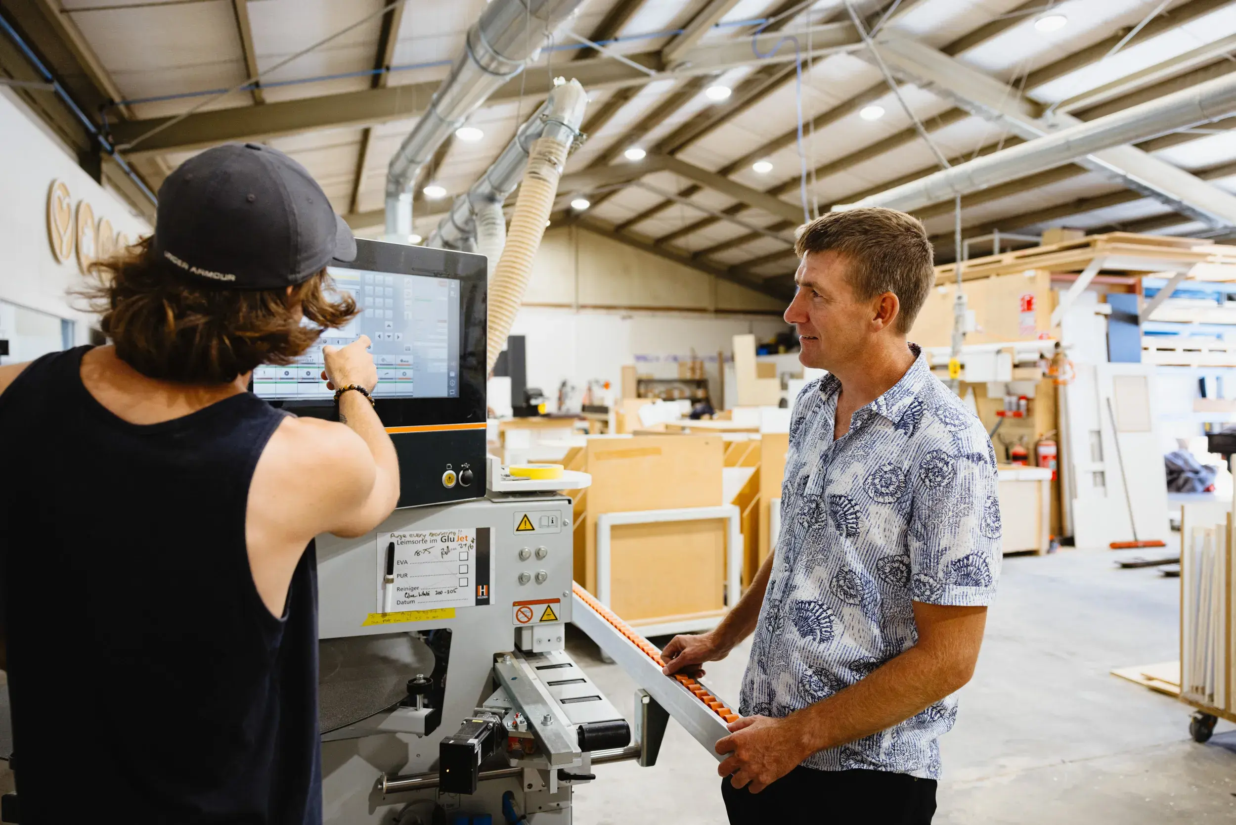 Two men working with a machine in a woodworking shop, one pointing at the screen and the other holding a piece of material